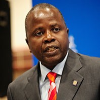 A man in a formal suit and striped tie speaks into a microphone. He stands against a blue background, conveying confidence and authority.