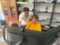 Tony sitting behind two computer monitors while another person gives instructions on the screen.