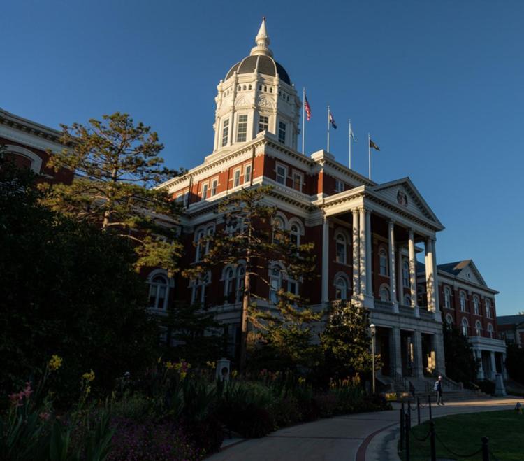 Historic red-brick building named Jesse Hall, with a white domed tower and columns, adorned with flags. Sunlit facade against clear blue sky, surrounded by trees.