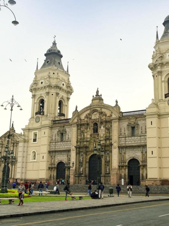 Historic cathedral facade with intricate stone carvings flanked by two towers against a clear sky. People walking and gathering nearby create a lively atmosphere.