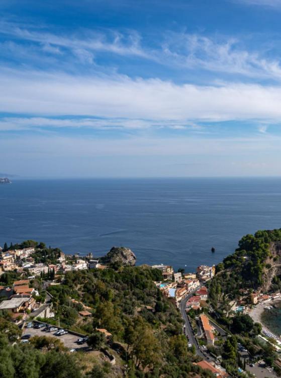 A coastal Italian town with red-roofed buildings and lush greenery overlooks the blue Mediterranean Sea. The sky is bright with scattered clouds.