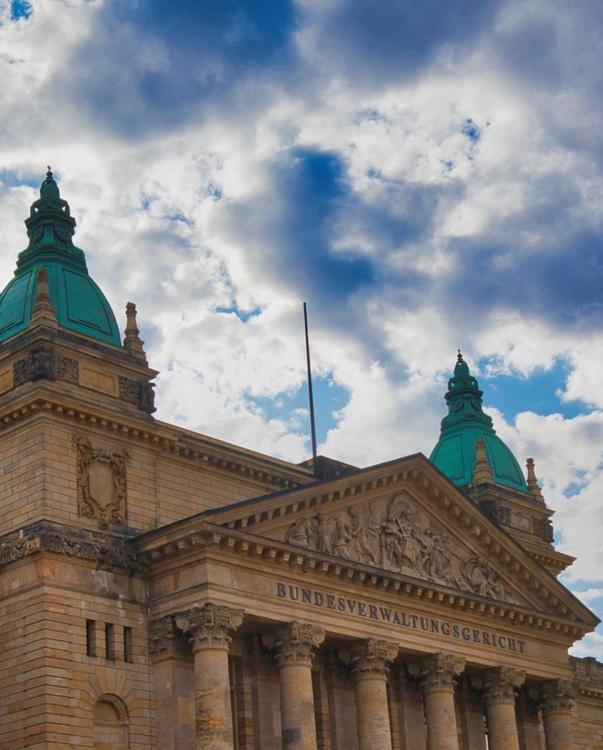 Historic stone building with green domes and ornate columns under a partly cloudy sky. Text on facade reads "Bundesverwaltungsgericht," conveying grandeur.