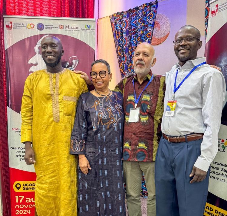 Four people stand together smiling, wearing colorful traditional and formal attire. 