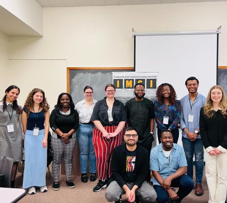 A group of eleven diverse individuals smiling for a photo in a classroom setting. A banner in the background reads "Interdisciplinary Migration Studies Institute."