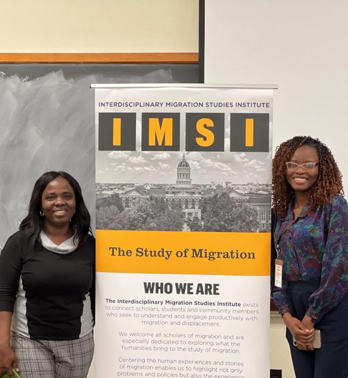 Two people stand smiling beside an IMSI banner for the Interdisciplinary Migration Studies Institute, in a classroom with a chalkboard backdrop.