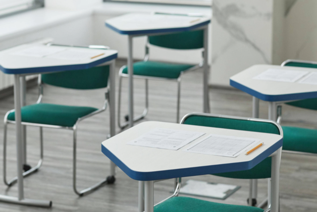 Empty classroom with modern desks and green chairs. Each desk has a sheet of paper and a pencil, evoking a quiet, studious atmosphere.