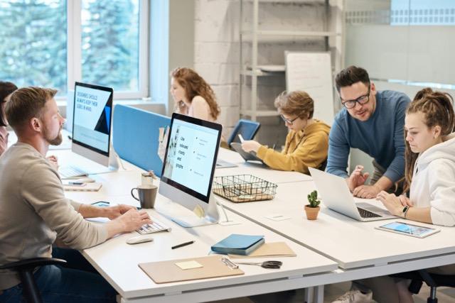 Open-plan office with six people working at desks. Two men collaborate near a laptop, others focus on desktop screens. Bright, collaborative atmosphere.