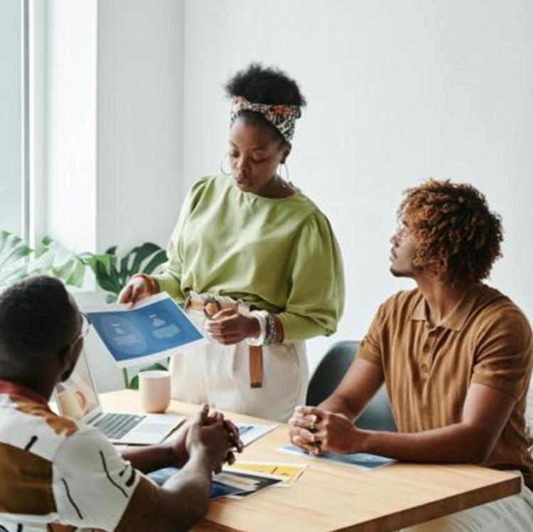 Three people in a meeting room. A woman holding a document stands, discussing with two seated men. The atmosphere is collaborative and focused.